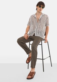 Kurzärmliges, knöpfbares Hemd mit gemustertem Design in Weiß-, Grau- und Rosatönen; kombiniert mit olivgrünen Hosen und braunen Sandalen.