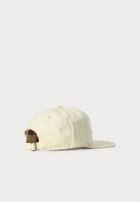 Gorra de béisbol ajustable en color beige con hebilla de metal, ojales visibles y visera plana, colocada sobre una superficie blanca.