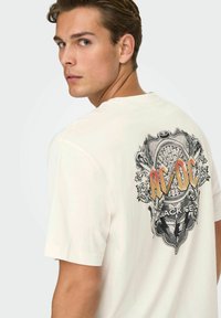 Cream-colored cotton t-shirt featuring a detailed black and orange graphic on the back, textured with ornate patterns and lettering.