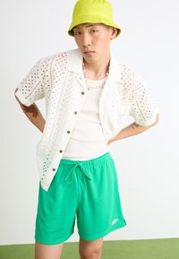 Cămașă albă cu model din dantelă, pantaloni scurți sport verzi cu șnur și o pălărie de tip bucket verde lime.