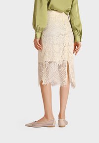 Gonna a matita in pizzo avorio con motivo floreale, spacchi laterali e orlo frangiato; abbinata a scarpe basse beige e una blusa verde.