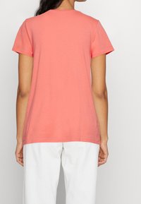 Koraliowy t-shirt z krótkim rękawem, okrągły dekolt, miękki materiał bawełniany, lekko dłuższy tył, zestawiony z białymi spodniami.