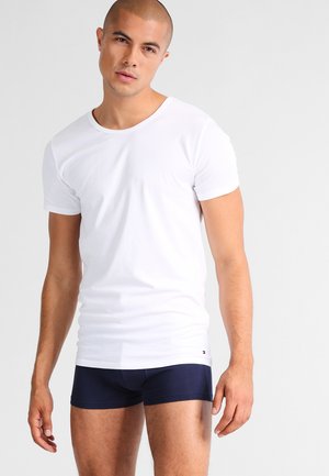 Man with shaved head wearing a plain white short-sleeve t-shirt and navy blue boxer briefs against a light background.