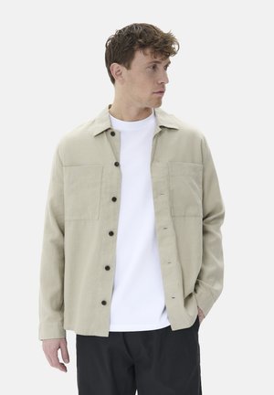 Beige button-up shirt made of lightweight fabric, featuring two chest pockets, black buttons, and a relaxed fit over a white T-shirt.