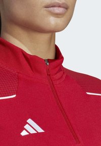 Giacca atletica rossa con colletto alto, chiusura con zip a metà, tessuto testurizzato e logo bianco del marchio. Presenta elementi a contrasto e una finitura liscia.
