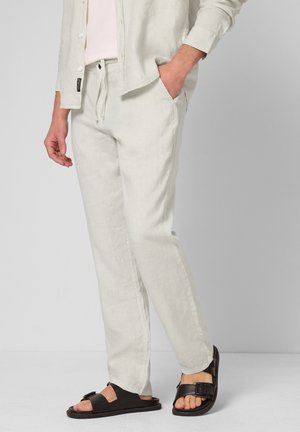 Person iført lyse beige hørbukser og en åbnet skjorte med den ene hånd i lommen, kombineret med sorte sandaler med spænde.