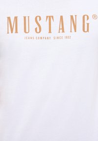 Weißes Baumwoll-T-Shirt mit großem orangefarbenem Schriftzug "MUSTANG" und dem Slogan "JEANS COMPANY SINCE 1932" darunter. Glatte Textur, kurze Ärmel.
