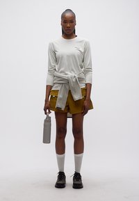 Γυναίκα που φορεί λευκό πουκάμισο με μακριά μανίκια, μουσταρδί σορτς, γκρι πουλόβερ δεμένο στη μέση, κρατώντας γκρι μπουκάλι νερού, στέκεται σε λευκό φόντο.