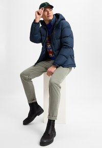 Navy pufferjack met capuchon, groene trui, olijfgroene broek en zwarte veterschoenen. Model zit op een voetstuk en past een pet aan.