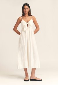 Valge midi-pikkusega kleit sõlmitud esiosa detailiga, spagetiribadega, tekstuurse kangaga ja lahtise lõikega. Kandmisel tasased tan toonis sandaalid.