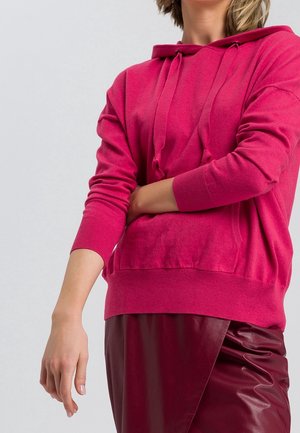 Osoba ubrana w jaskraworóżową bluzę z kapturem i sznurkami oraz ciemnordzoną skórzaną spódnicę, z jedną ręką skrzyżowaną, a drugą opuszczoną.