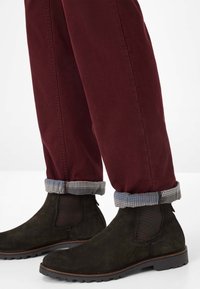 Stivali Chelsea in camoscio verde scuro con una superficie texturizzata, abbinati a pantaloni bordeaux. I polsini presentano un bordo a motivi blu e bianco.