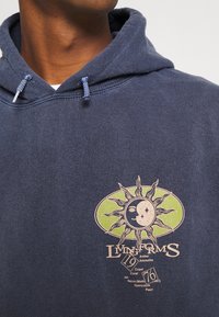 Sudadera de algodón azul marino con un gráfico de sol y luna en tonos beige claro y verde. Incluye cordones con extremos texturizados para ajustarla.