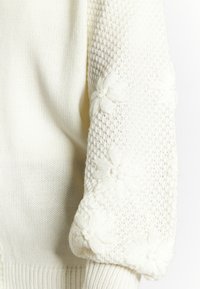 Weißer Strickpullover mit strukturierten, gehäkelten Ärmeln und floralen Details. Weicher Stoff mit einer Mischung aus offenen und geschlossenen Maschenmustern.