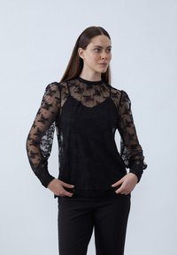 Black sheer blouse with floral lace pattern, high neckline, puffed sleeves, and fitted inner layer; worn with black trousers.