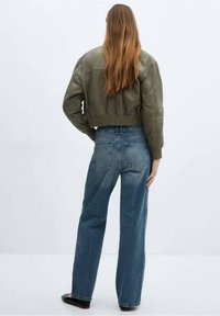 Mujer con cabello largo castaño que lleva una chaqueta corta verde oliva, jeans rectos azul y zapatos negros, de pie frente a una pared blanca.