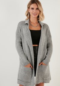 Cárdigan de punto gris oversize con cuello, textura acanalada y bolsillos delanteros. Diseño de frente abierto con mangas largas.