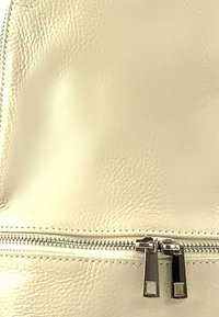 Crème leren tas met een zichtbare rits aan de onderkant, voorzien van metalen hardware en een subtiele textuur. Stiksel details langs de rand.