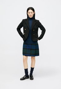 Črni dvoredni blazer z zlatimi gumbi, mornarsko modra pletena jopica, karirana krilo v modro-zelenih odtenkih, črni loaferji in mornarsko modre nogavice.