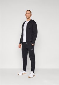 Čierna mikina na zips s vreckami, nosená cez biele tričko, doplnená čiernymi teplákmi s textovým detailom a bielymi teniskami s hnedými podrážkami.