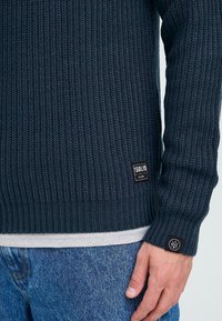 Close-up of a person wearing a dark navy knit sweater over a grey shirt and blue jeans, with brand tags visible on the sweater.