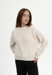 Beige Kabelstrickpullover mit grober Textur, Rautenmuster und gerippten Bündchen. Lässige Passform und runder Halsausschnitt. Getragen mit schwarzen Hosen.