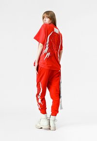 Una camiseta roja y joggers a juego presentan diseños de alas en blanco y plateado en la parte posterior y lateral, fabricados con una tela suave. Botines blancos de tobillo completan el look.