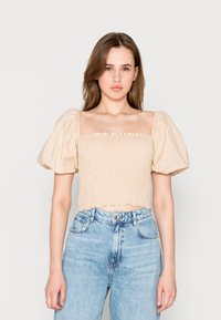 Top court à manches bouffantes beige clair avec une texture froncée et des bordures en volants, associé à un jean bleu taille haute.