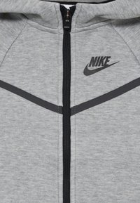 Sivi zip-up dukserica od mekog materijala s crnim patentnim zatvaračem, s crnim Nike logotipom i tamnosivim naglasnim linijama na ramenima.