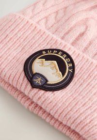Roze gebreide muts met geribbelde textuur. Voorzien van een zwart, rond embleem met een bergontwerp en het woord "SUPERDRY" geborduurd op het embleem.