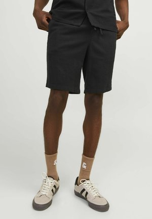Schwarze Shorts aus strukturiertem Stoff, knielang, mit elastischem Bund und Kordelzug. Kombiniert mit beigen Sneakers und braunen Socken.