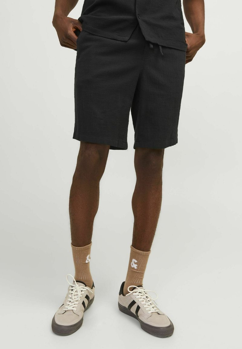 Shorts noirs en tissu texturé, longueur genoux, taille élastique avec cordon de serrage. Assortis à des baskets beiges et des chaussettes brun clair.