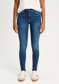Jean skinny en denim bleu foncé avec une texture lisse, présentant un design à cinq poches traditionnel et une fermeture éclair. Porté avec des baskets blanches.