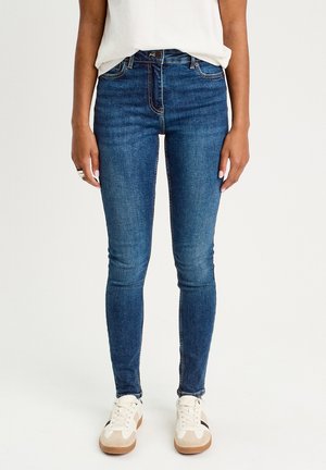 Schmale Jeans aus dunkelblauem Denim mit glatter Textur, ausgestattet mit einem klassischen Fünf-Taschen-Design und einem Reißverschluss. Getragen mit weißen Sneakers.