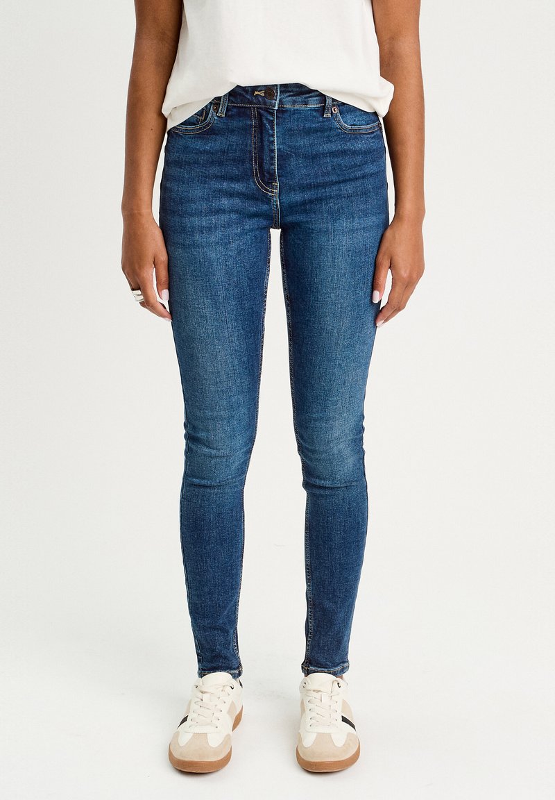 Jean skinny en denim bleu foncé avec une texture lisse, présentant un design à cinq poches traditionnel et une fermeture éclair. Porté avec des baskets blanches.