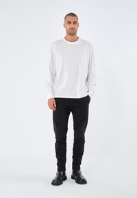 Witte longsleeve t-shirt, zwarte slim-fit broek en zwarte enkellaarzen. Gladde stof en eenvoudig ontwerp, staand tegen een effen achtergrond.