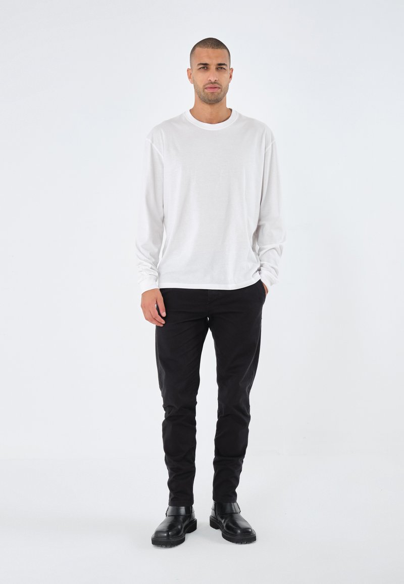 Witte longsleeve t-shirt, zwarte slim-fit broek en zwarte enkellaarzen. Gladde stof en eenvoudig ontwerp, staand tegen een effen achtergrond.