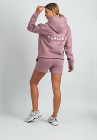 Világos lila kapucnis pulóver logóval a hátán, hozzáillő rövidnadrággal. Az anyag puhanak tűnik, laza szabással.