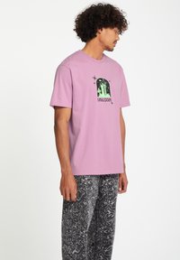 Lila katoenen t-shirt met korte mouwen, met een zwarte grafische print en groene accenten. Gecombineerd met grijze met patroon broek.