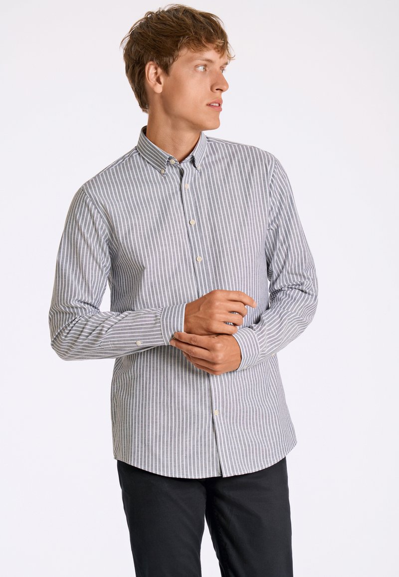 Button-up shirt in blue and white stripes, made of smooth fabric with a collar, long sleeves, and rounded hem. Black pants are worn.