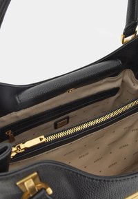 Sisäpuoli mustasta nahkaisesta käsilaukusta, jossa on beige kangasvuori, johon on painettu "GUESS", musta vetoketjusalkku ja kultainen vetoketjukiinnitys.