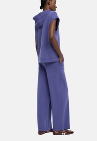 Kobieta z warkoczami w fioletowej bluzie bez rękawów i szerokich spodni matching, stojąca z rękami w kieszeniach, w brązowych butach z guzikami.