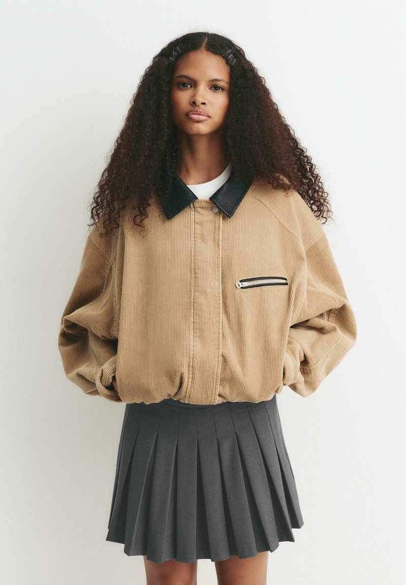 Jeune femme avec des cheveux bouclés portant une veste en velours côtelé beige avec un col noir et une jupe plissée noire, se tenant devant un fond blanc.