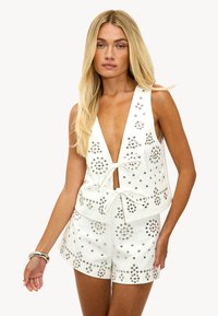 Top blanco sin mangas con un escote en V profundo, adornado con tachuelas plateadas y un lazo en la parte delantera. Combinado con pantalones cortos a juego decorados con tachuelas similares.