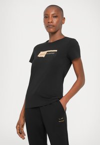 Tricou negru din bumbac, cu un grafic bej ce include textul "Armani Exchange". Mâneci scurte și decolteu rotund, asortat cu pantaloni negri.