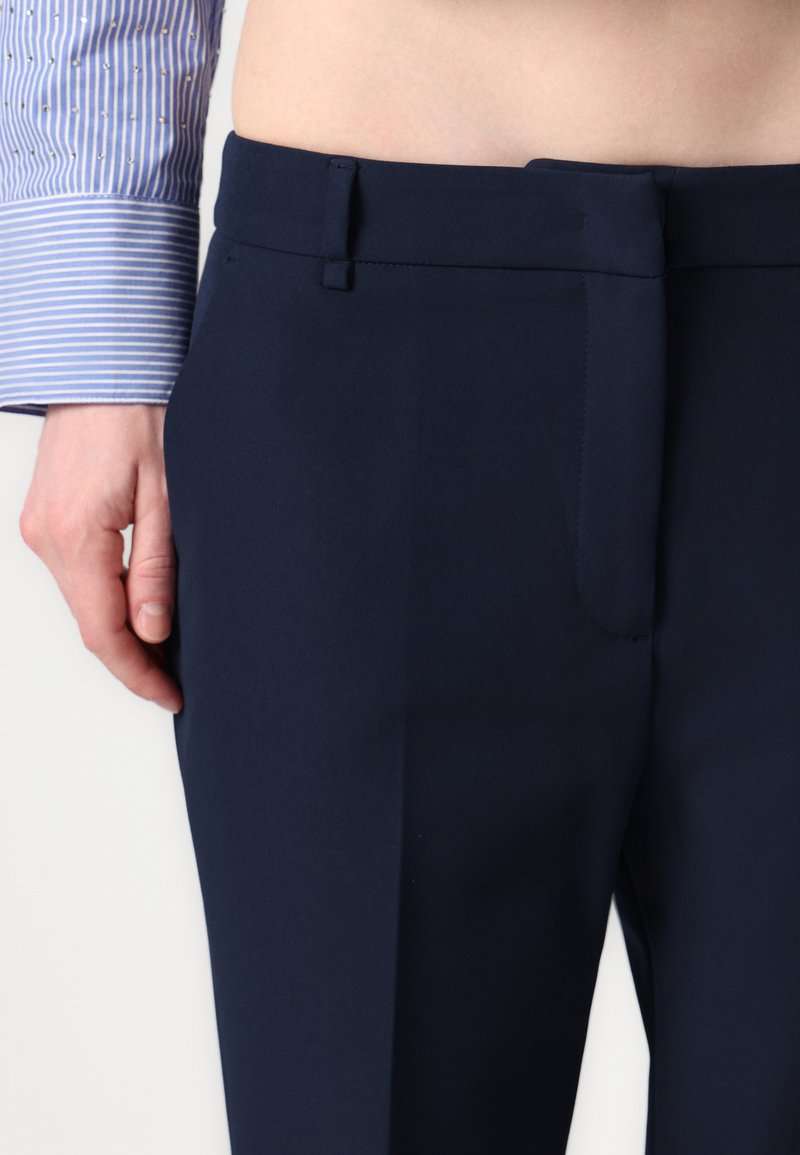 Pantaloni tailored blu navy con una texture liscia, frontale piatto e una tasca posteriore singola. Abbinati a una camicia a maniche lunghe a righe blu chiaro.