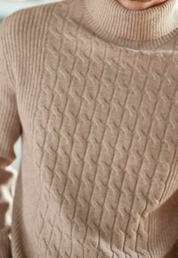 Suéter beige tejido con un patrón texturizado de trenzas y un cuello alto acanalado. La tela parece suave con un acabado acogedor.