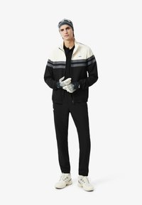 Giacca zip-up bianca e nera con strisce grigie, indossata sopra una polo nera e pantaloni neri abbinati. Gli accessori comprendono guanti e cappello grigi.
