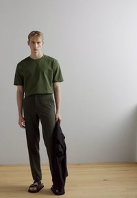 T-shirt de algodão verde e calças ajustadas combinadas; textura suave, corte descontraído; modelo segura uma jaqueta preta; chão de madeira e fundo claro.