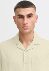 Chemise à manches courtes vert clair en tissu texturé, avec un col échancré, fermeture à boutons sur le devant et poche sur la poitrine gauche.
